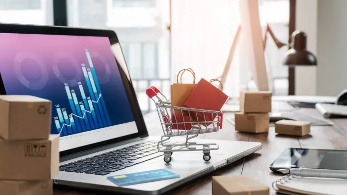 Laptop showing a growth chart beside a small shopping cart with shopping bags and cardboard boxes