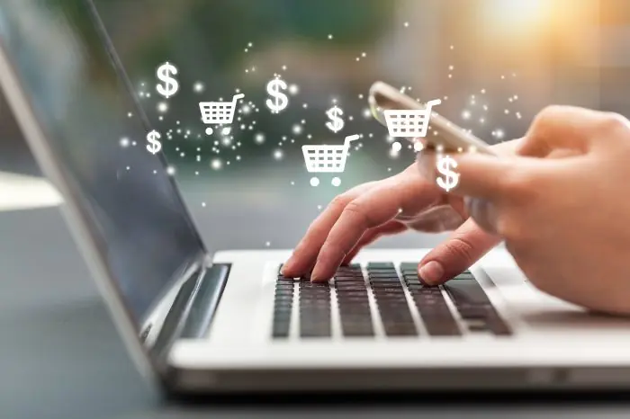 Hand using a laptop and smartphone with shopping cart and dollar icons floating above, representing online sales
