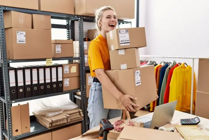 Smiling woman holding a stack of shipping boxes in a small e-commerce stockroom with a laptop on the desk