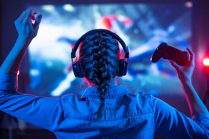 Woman seen from behind wearing headphones and holding a game controller, facing a large screen