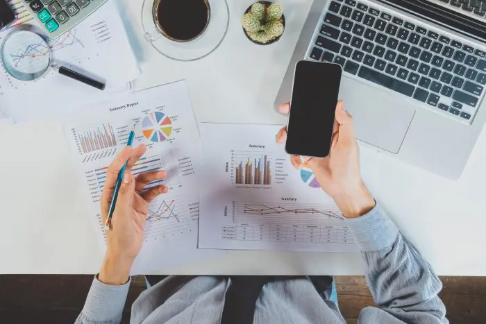 Hands holding a smartphone and pen over printed analytics reports and charts on a desk