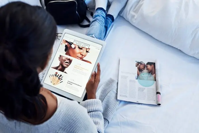 Woman on a bed viewing a beauty blog on a tablet beside an open magazine
