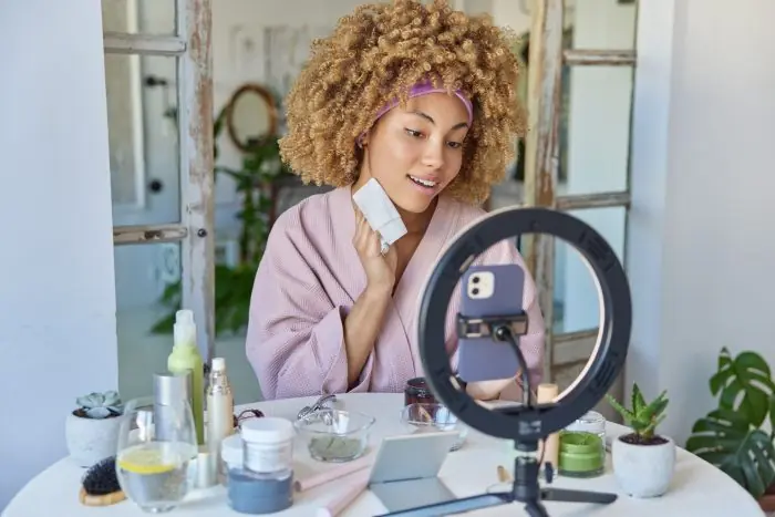 Woman in a robe holding a skincare tool and smiling at a phone on a ring light while filming beauty content
