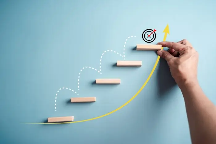 Hand placing a wooden step toward a target on a rising yellow arrow, representing growth marketing