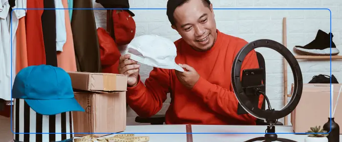Smiling man in an orange sweater holding a white cap in front of a ring light, filming a product video
