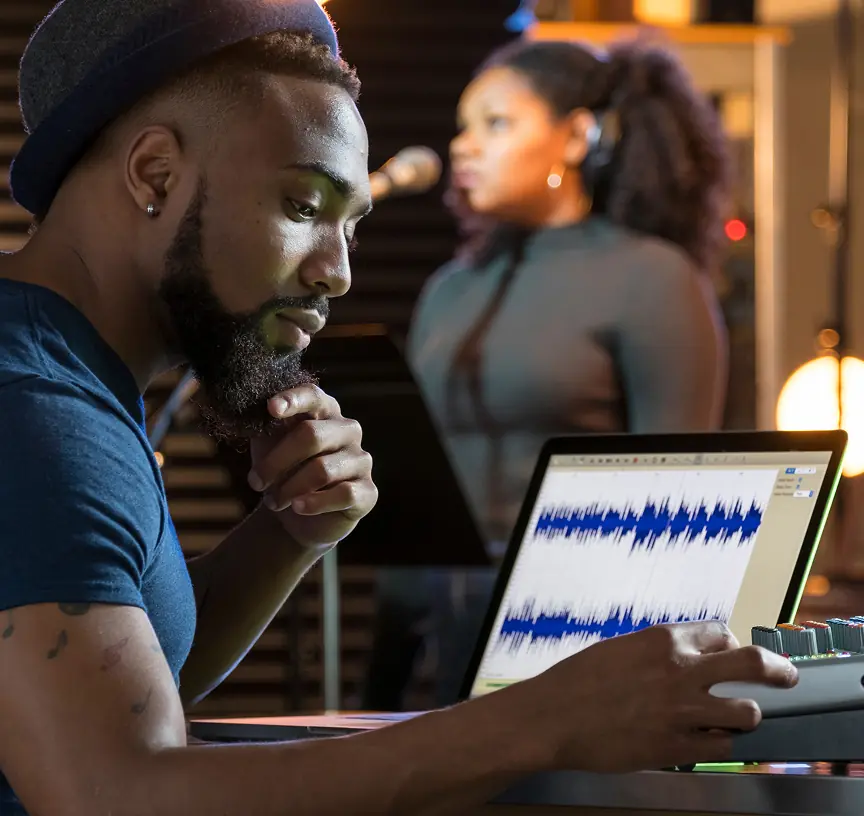 Music producer at a laptop showing an audio waveform, with a singer behind him in a recording studio